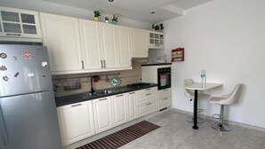A modern kitchen with white cabinets, a stainless steel refrigerator, a gas stove, and a built-in oven. There are two white bar stools and a small table with a water bottle.