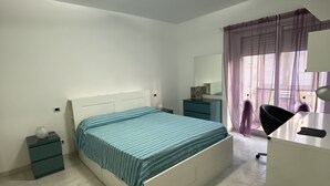 A bedroom with a bed, a desk, a chair, a nightstand, a lamp, and a window with curtains.