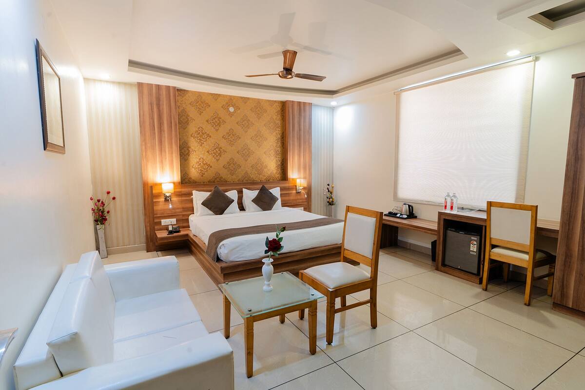 A hotel room with a bed, a desk, a chair, a small table, a ceiling fan, and a window with blinds.