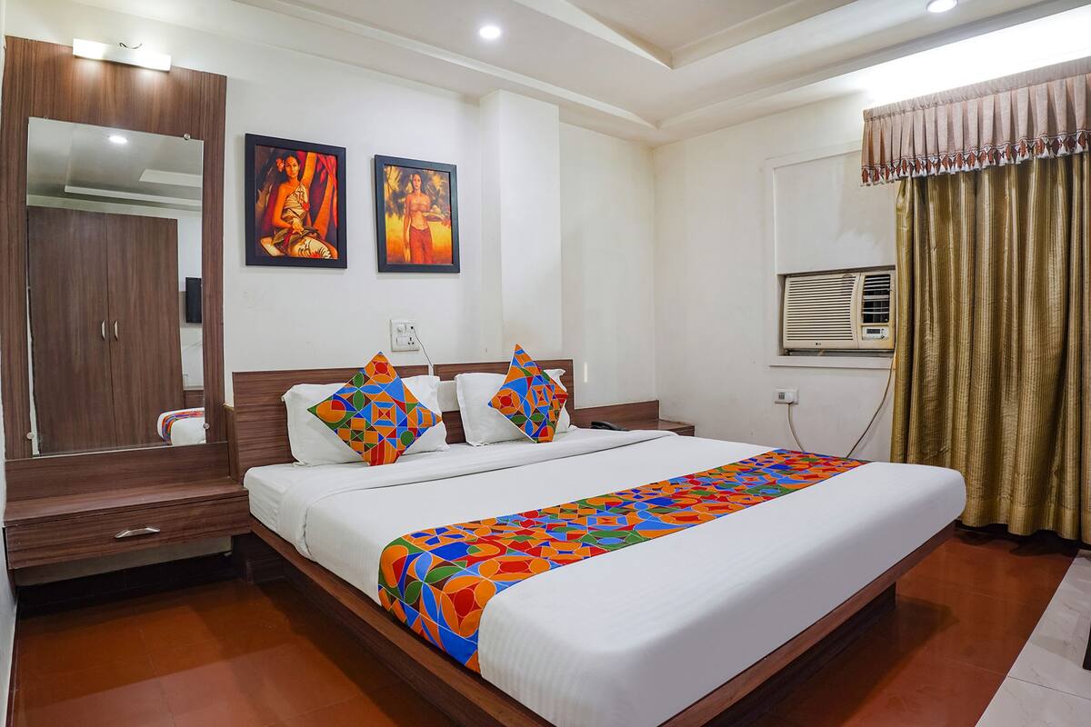 A hotel room with a large bed, wooden wardrobe, two wall-mounted paintings, an air conditioning unit, and a curtain.