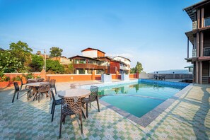 A swimming pool with a tiled deck, surrounded by outdoor furniture and a building with multiple balconies. - Treebo Seven Days, Mahabaleshwar (Mahabaleshwar)