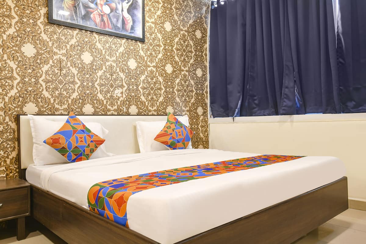 A neatly made bed with white linens and colorful pillows, a patterned wallpaper, and a window with dark curtains.
