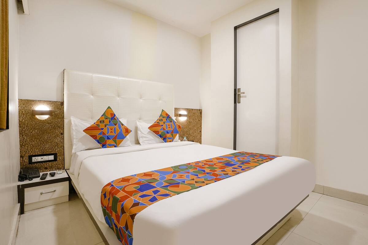 A hotel room with a large bed, white bedding, a colorful geometric throw blanket, and a nightstand with a phone and remote controls.