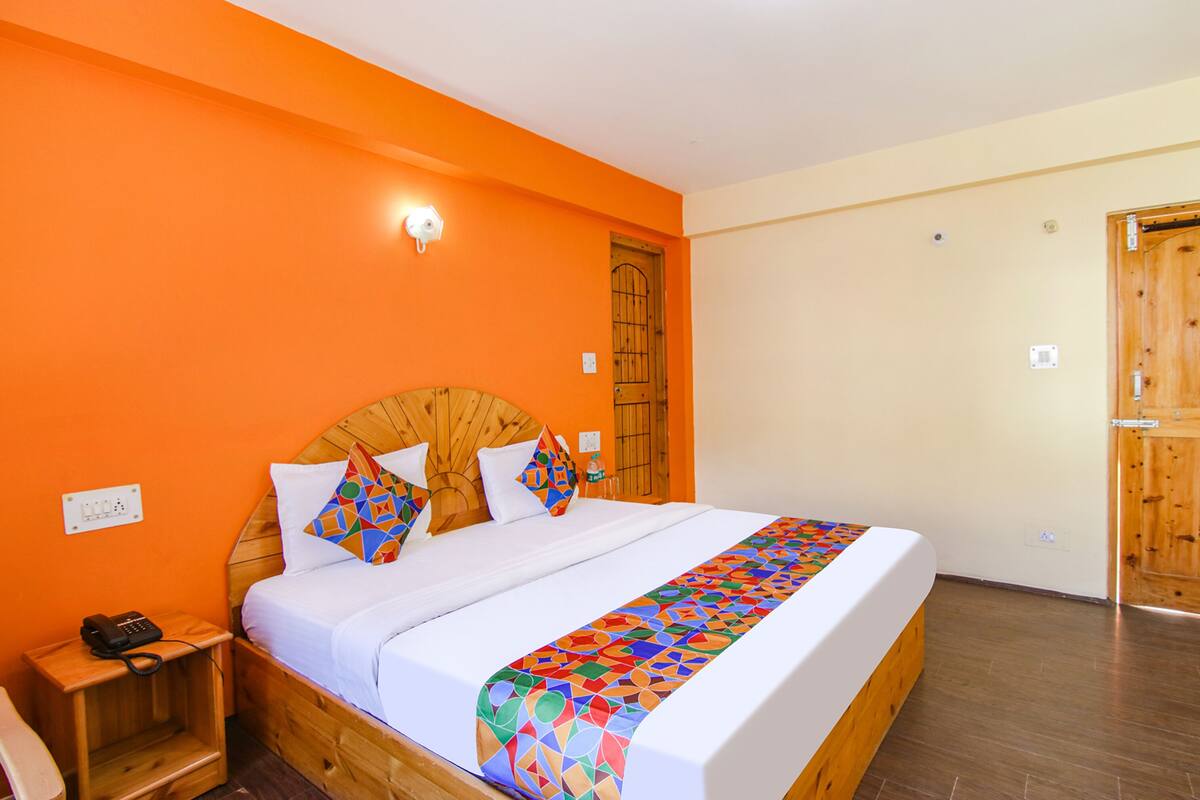 Ein Hotelzimmer mit orangefarbener Wand, einem hölzernen Kopfteil, einem Bett mit weißer Bettwäsche und bunten Kissen, einem Nachttisch mit Telefon und einer Holztür.