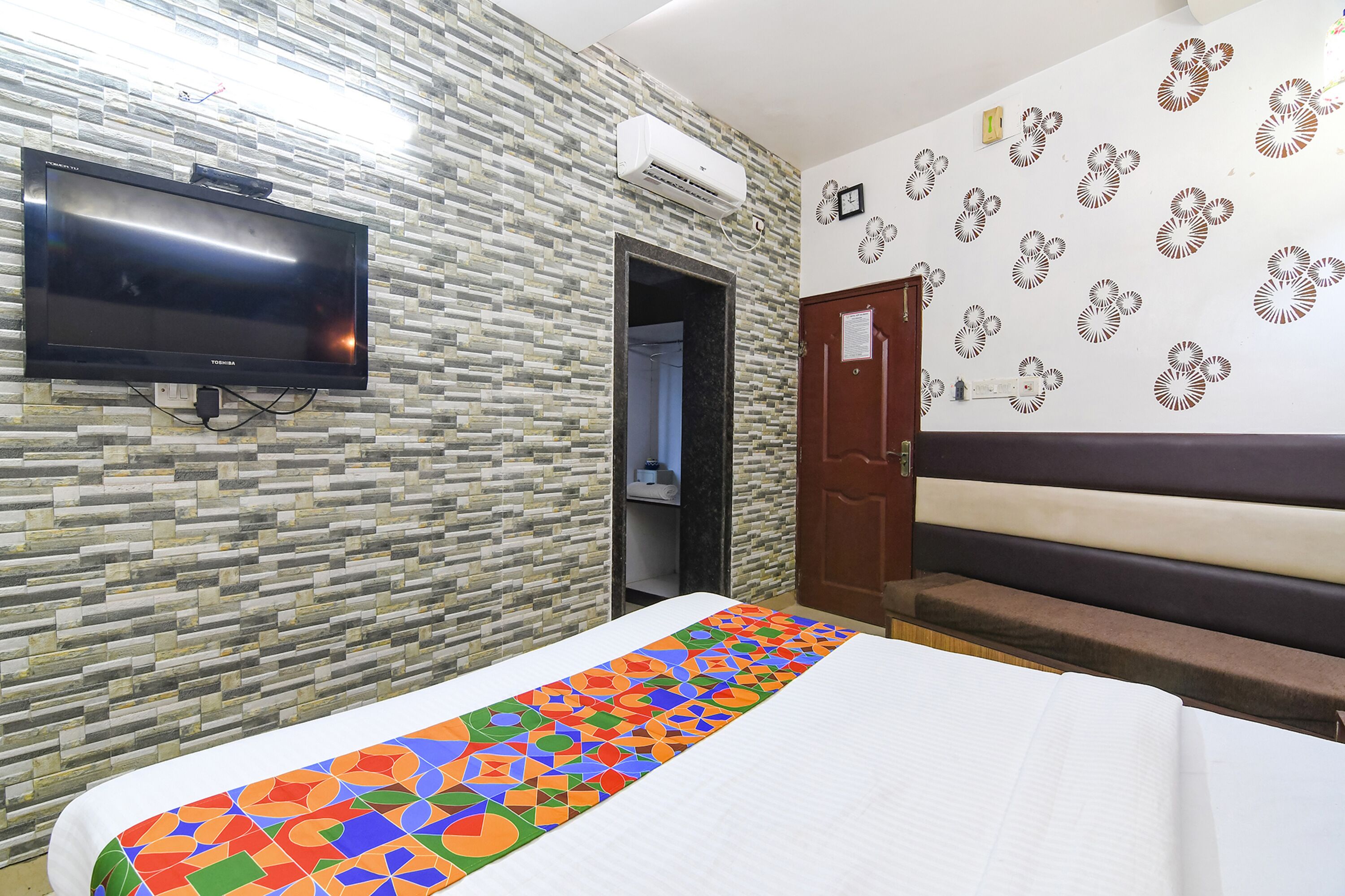 Une chambre d’hôtel avec un lit, un téléviseur fixé au mur, un mur à motifs, une porte et un banc.