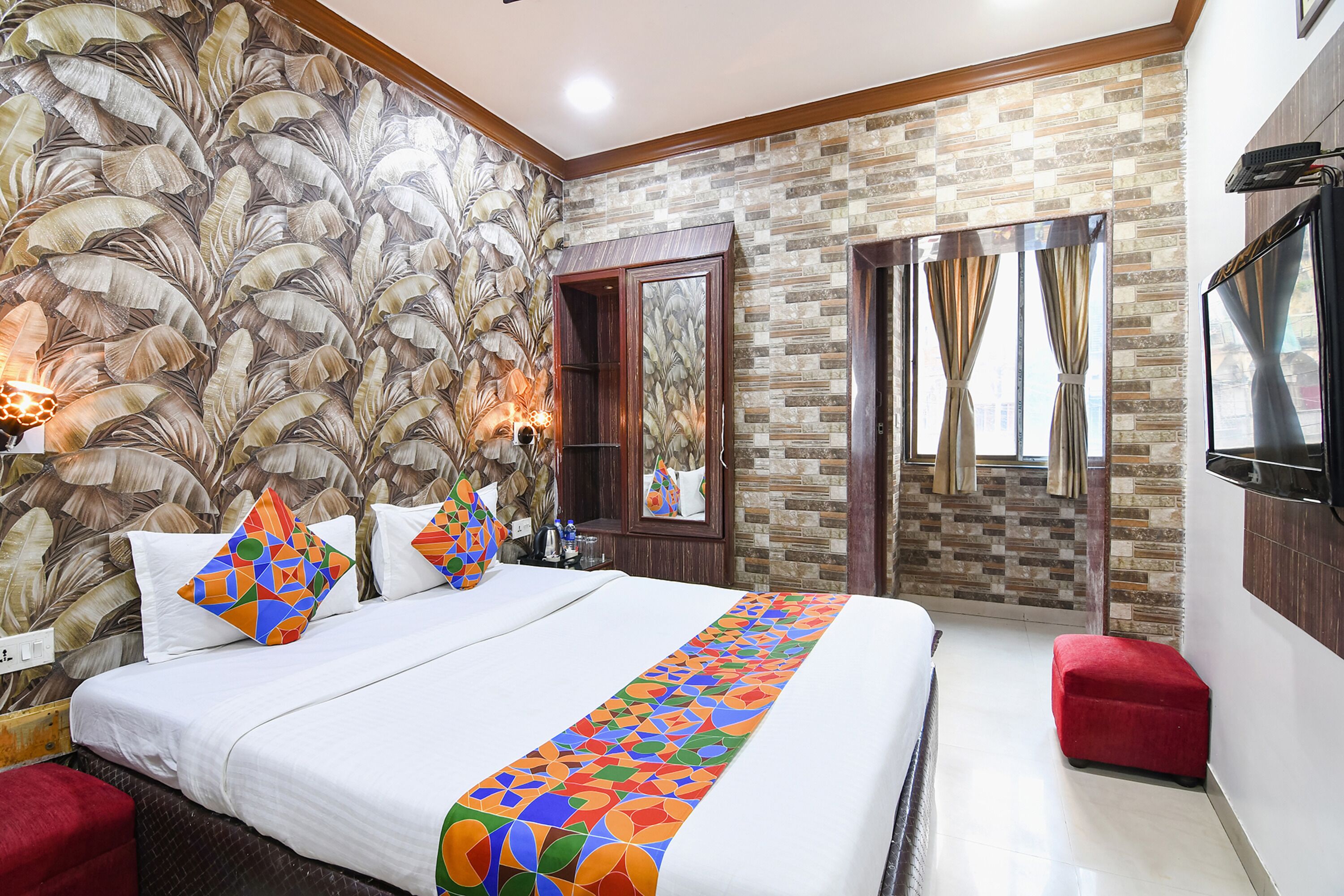 A hotel room with a large bed, a red ottoman, a mirror, and a window with curtains.
