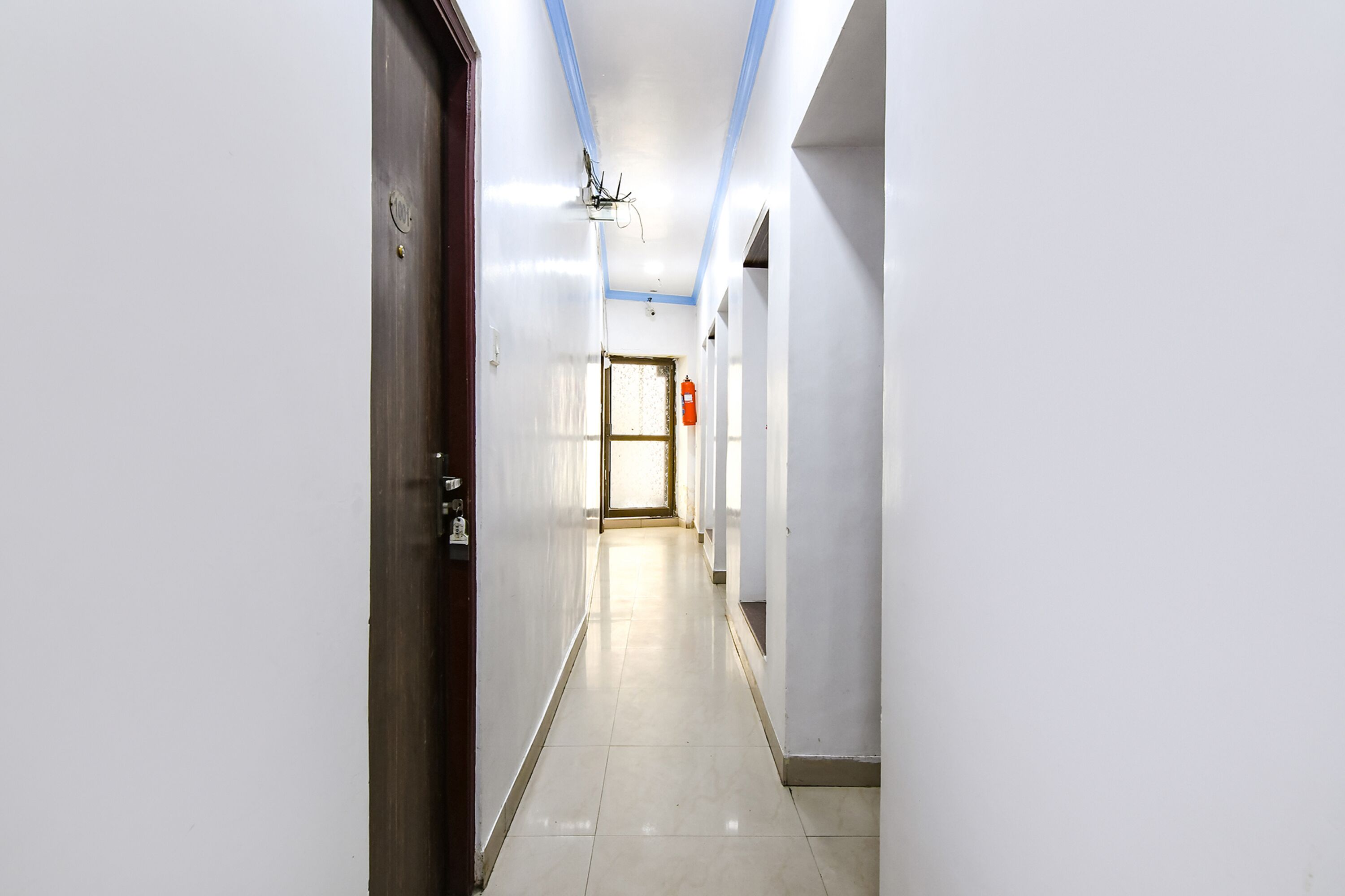 A hallway with a door on the left, a fire extinguisher on the right, and a ceiling light fixture.
