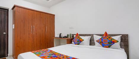 Una habitación de hotel con un armario de madera, una cama con colcha geométrica colorida y una mesita de noche con una lámpara y un cactus.
