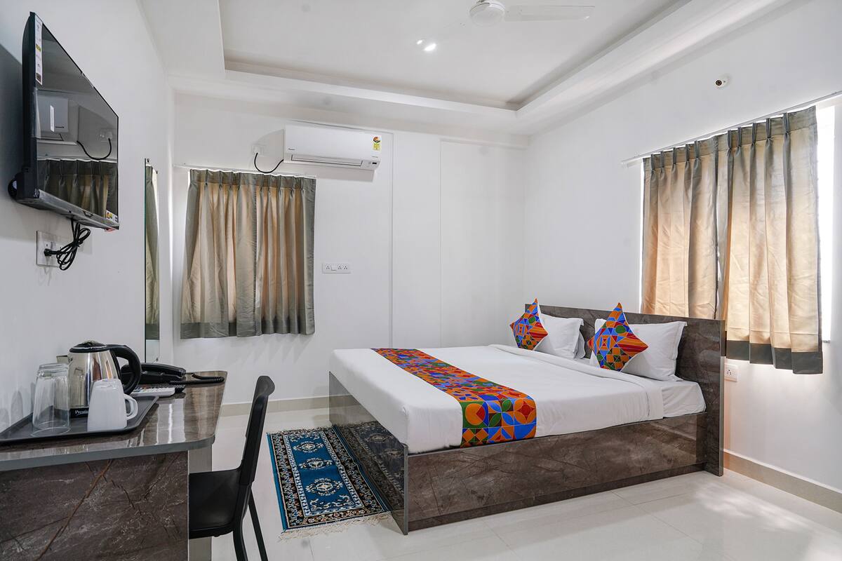 A hotel room with a bed, a desk with a telephone and a kettle, an air conditioning unit, and curtains.
