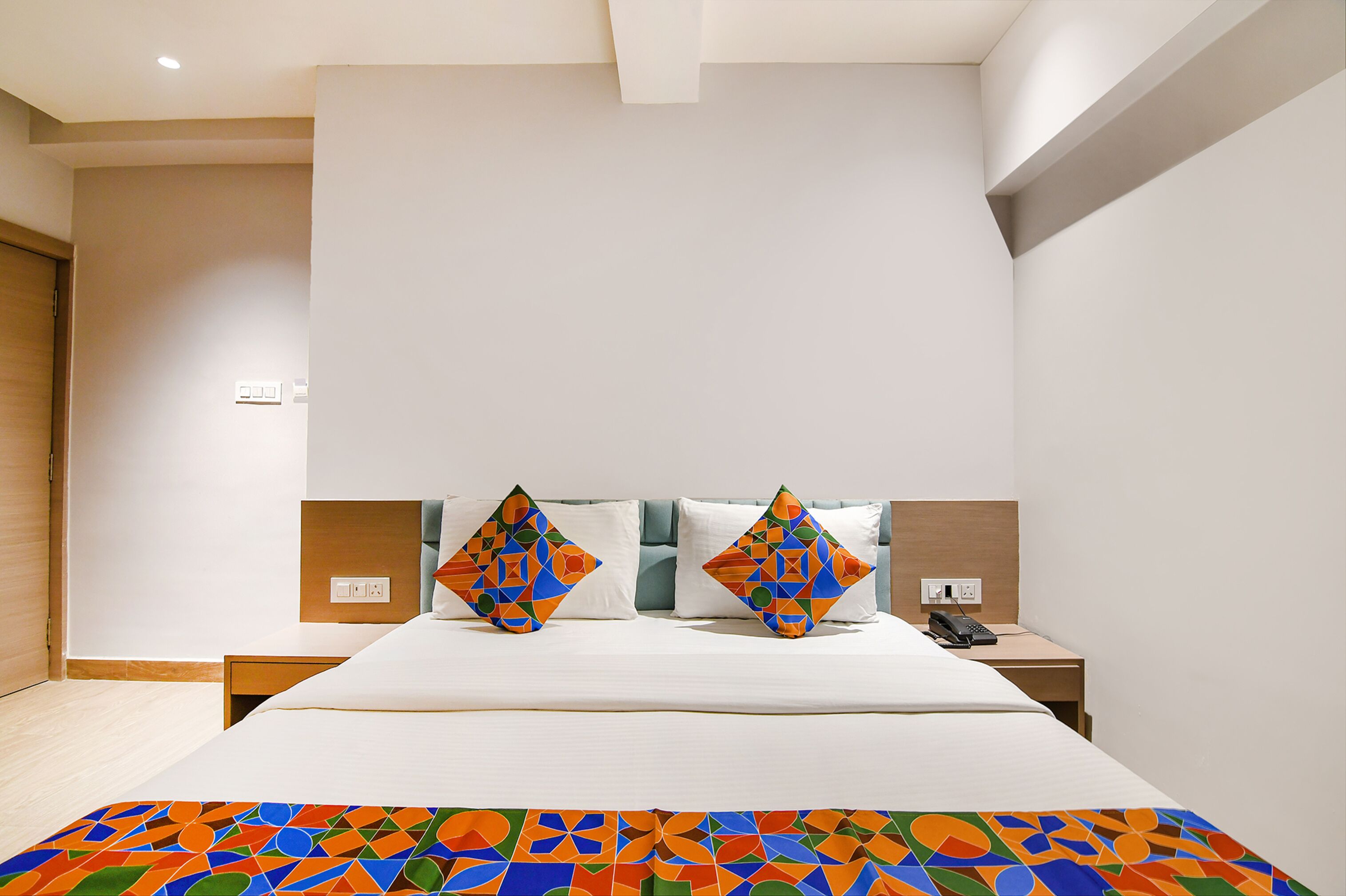 Quarto de hotel moderno com uma cama grande, dois travesseiros com padrões geométricos, uma mesinha de cabeceira com telefone e um cobertor colorido.