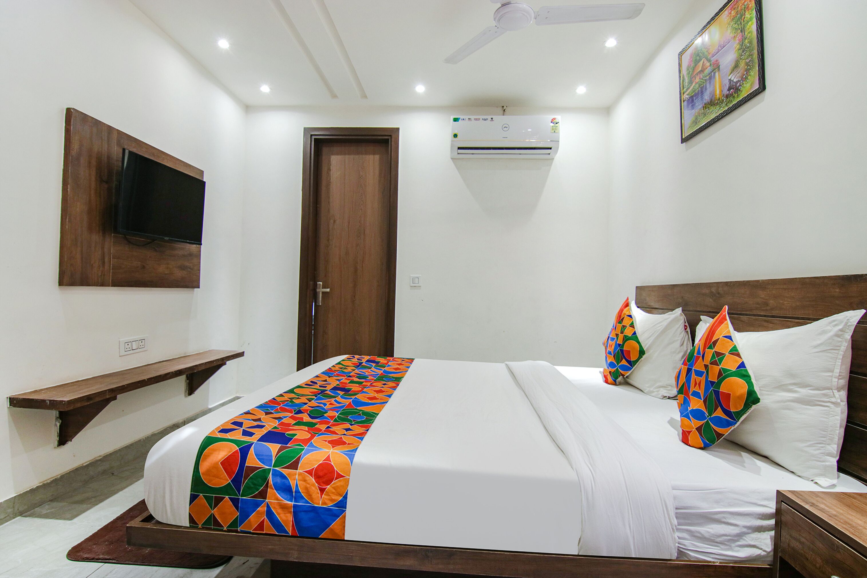 A hotel room with a bed, a TV mounted on the wall, an air conditioning unit, a wooden headboard, and a painting on the wall.