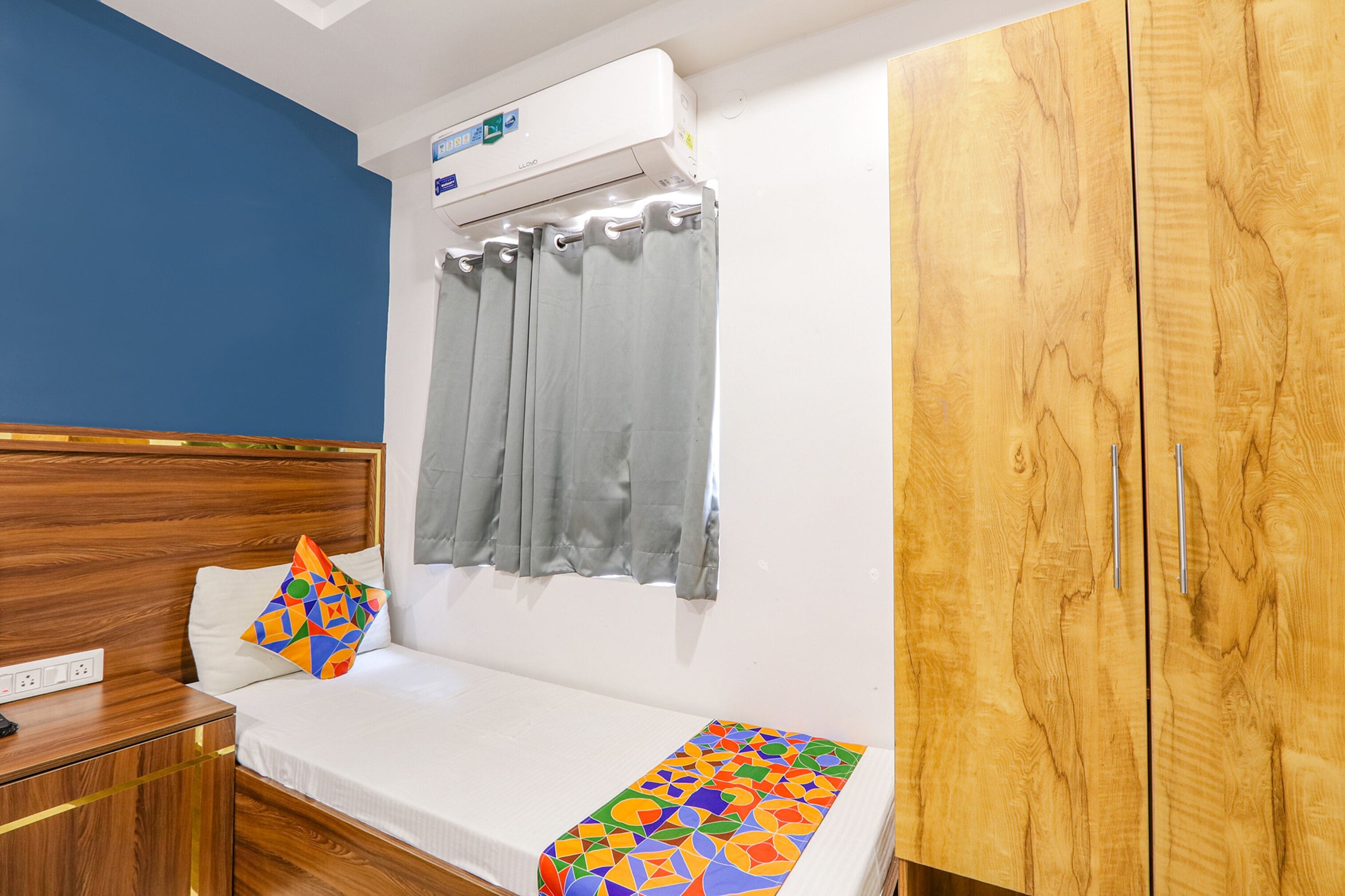 A hotel room with a wooden headboard, a bed with a colorful geometric-patterned bedspread, a gray curtain, and a wooden wardrobe.