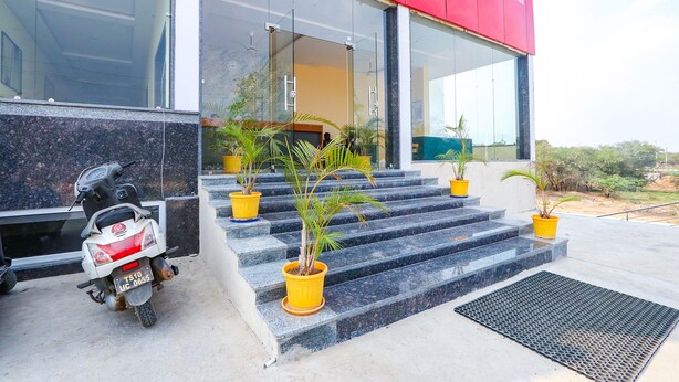 A modern building with a red facade, a glass entrance, and a set of steps leading to a door. There are potted plants on the steps and a scooter parked to the side.