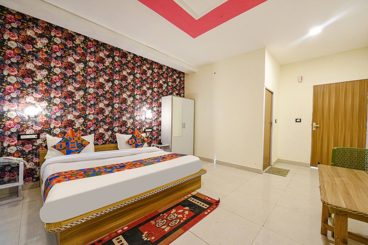 A bedroom with a floral wallpaper, a bed with a patterned bedspread, a wooden nightstand, a green chair, and a wooden door.