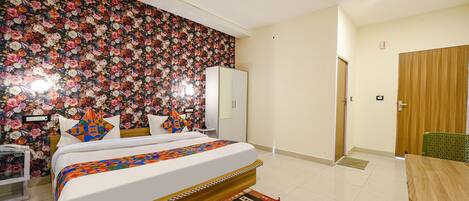 Un dormitorio con papel tapiz floral, una cama con colcha estampada, una mesita de noche de madera, una silla verde y una puerta de madera.