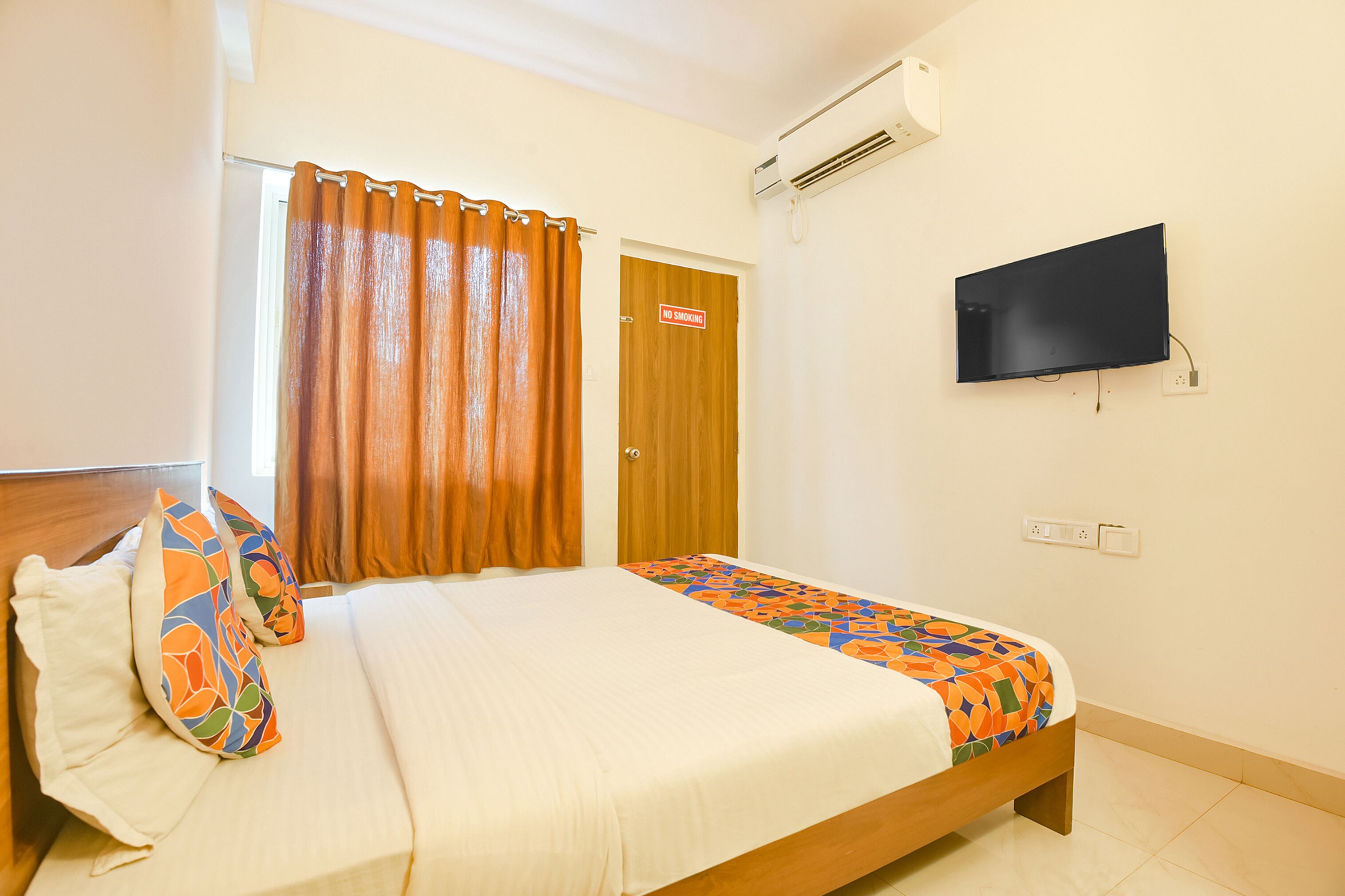 A hotel room with a bed, a TV mounted on the wall, a door with a "No Smoking" sign, and an air conditioning unit.