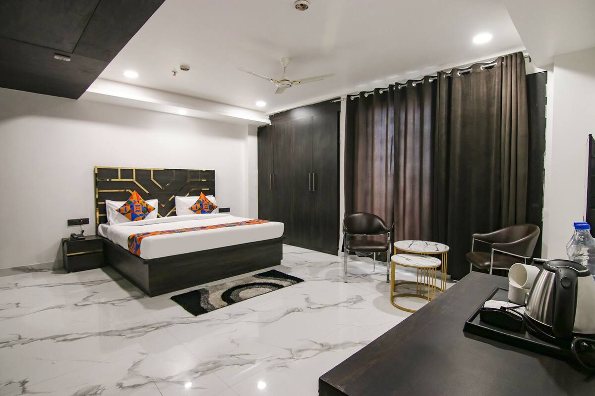 A modern hotel room with a large bed, dark wood wardrobe, a desk with a kettle, and a chair.
