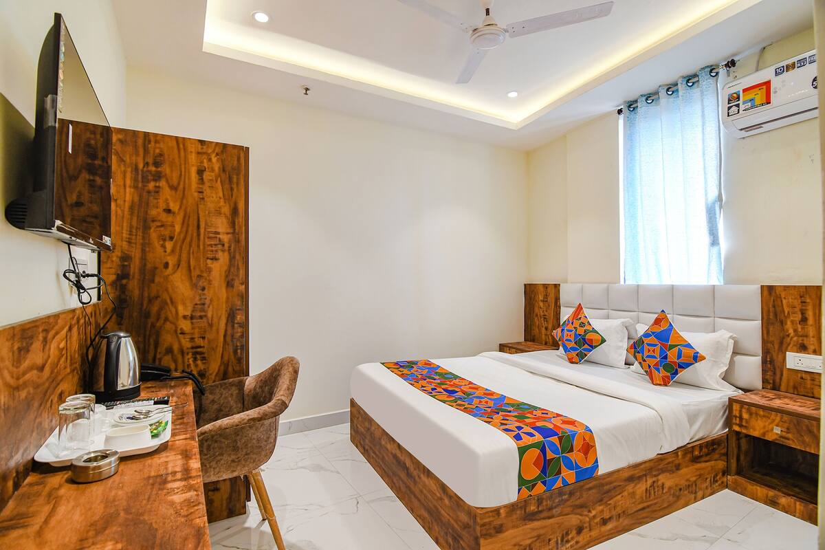 A bedroom with a wooden headboard, a bed with colorful pillows, a desk with a kettle, and a TV mounted on the wall.