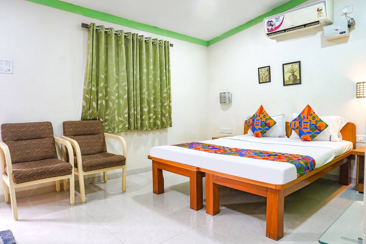 A hotel room with a bed, two chairs, a window with green curtains, and an air conditioning unit on the wall.