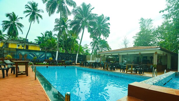A swimming pool with lounge chairs and tables, surrounded by palm trees and a building with a mural. - Fabhotel Grand Royal Palms (Benaulim)