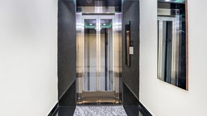 A modern elevator with a metallic finish and glass panels, located in a hallway with a marble floor and a mirrored wall. - Fabhotel Prime Ravila Grand (Hyderabad)