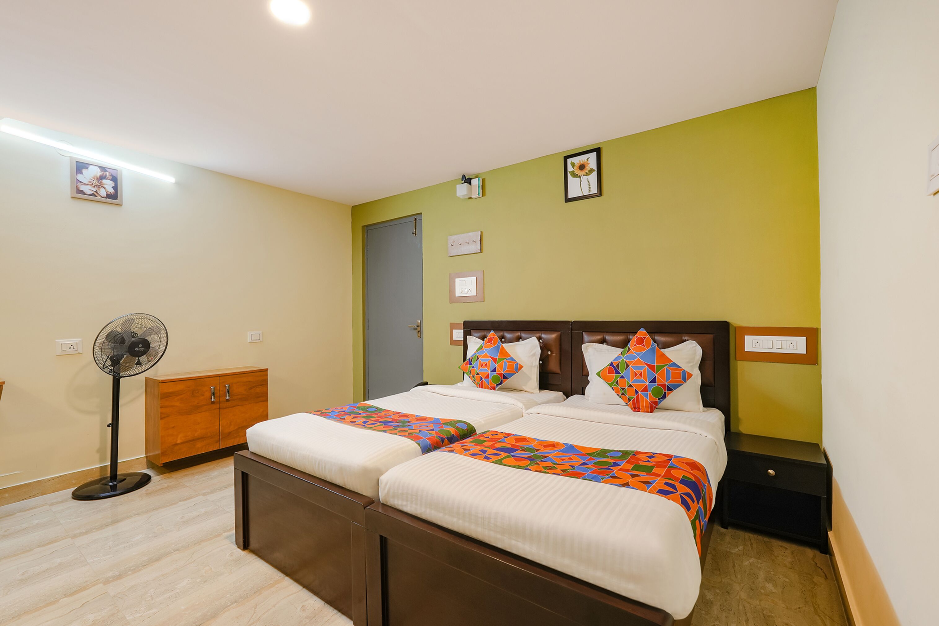 A hotel room with two beds, a wooden wardrobe, a standing fan, and a small nightstand.