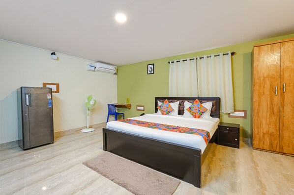 Un dormitorio con cama, armario, escritorio, silla, ventilador y refrigerador.