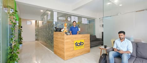 Zona de recepción con un mostrador de madera con la etiqueta 'fab hotels', una recepcionista, un sofá y una planta.