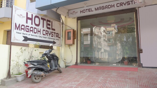 La entrada del Hotel Magadh Crystal, con un letrero visible, una moto estacionada y una planta en maceta.