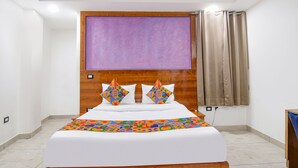 Une chambre à coucher avec une tête de lit en bois, un grand lit avec une literie blanche, deux coussins décoratifs, une couvre-lit colorée, une grande fenêtre avec des rideaux beige et un téléviseur fixé au mur.