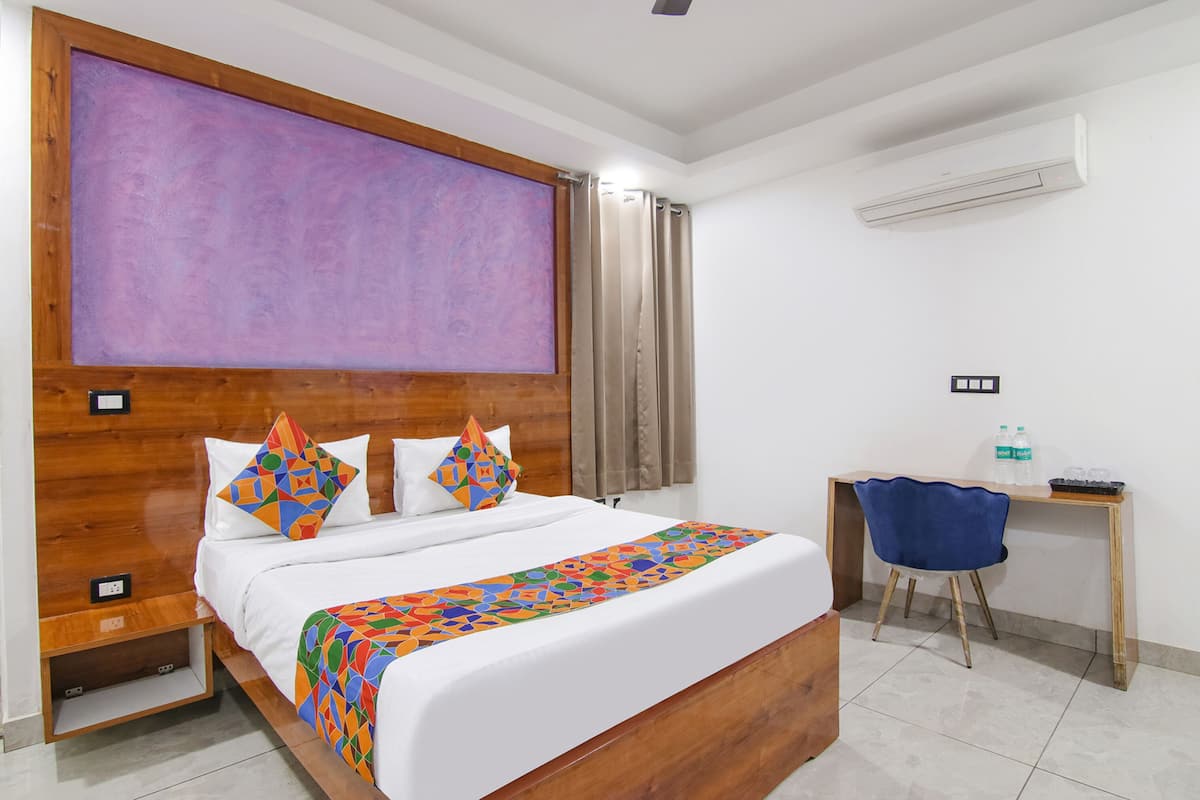 A hotel room with a wooden headboard, a large bed with colorful bedding, a desk with a chair, and an air conditioning unit.