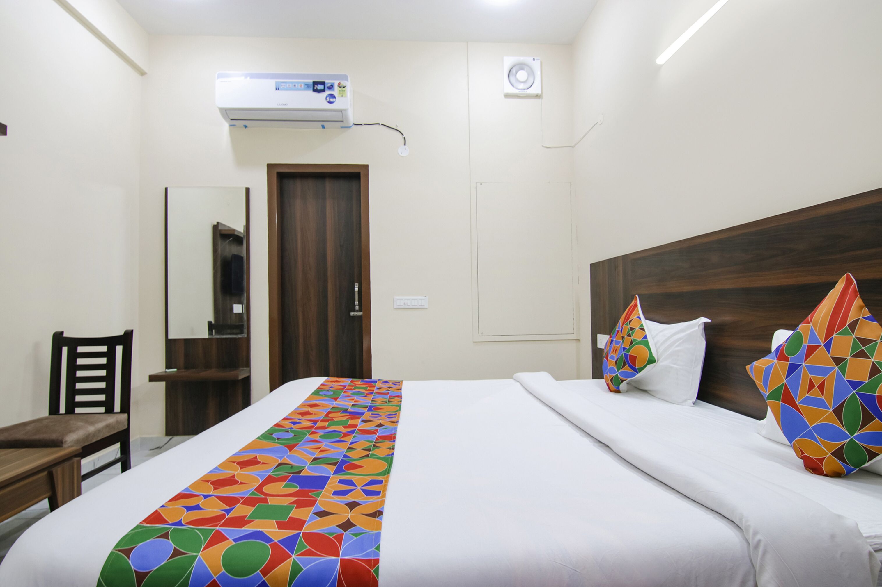 A hotel room with a bed, a desk, a chair, a mirror, and an air conditioning unit.