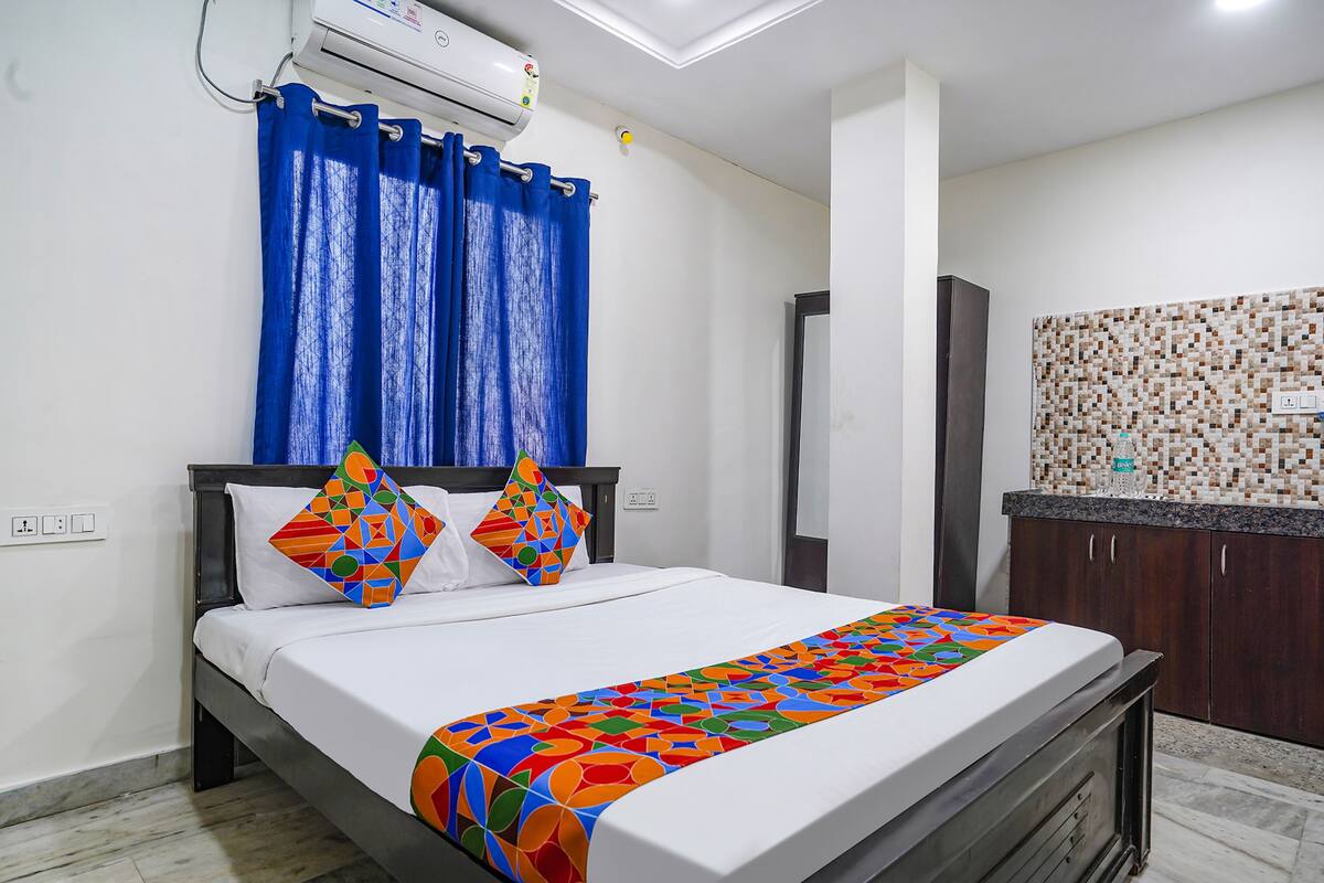 Cama bem arrumada com colcha geométrica colorida, cortina azul e ar condicionado fixado na parede.
