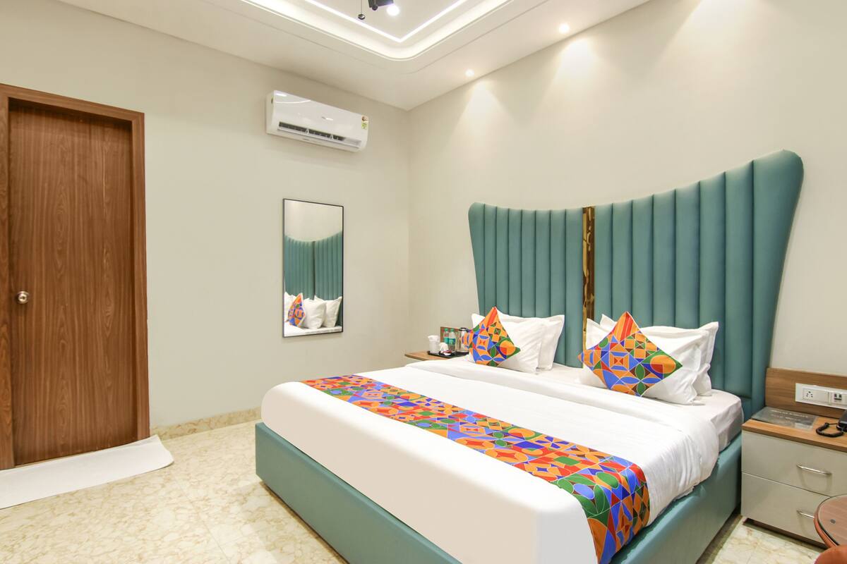 A hotel room with a large bed, a wooden door, a wall-mounted air conditioner, a bedside table with a phone, and a mirror.