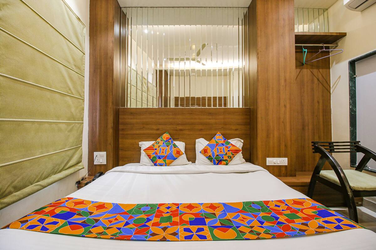 Une chambre d’hôtel avec un lit, deux oreillers à motifs géométriques, une couvre-lit colorée, une tête de lit en bois, une chaise et une fenêtre avec des stores.