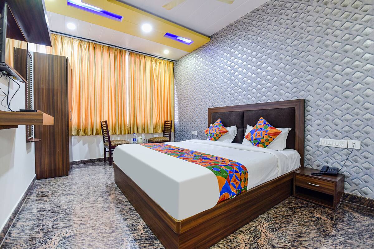 A hotel room with a large bed, wooden headboard, and a colorful bedspread. There is a desk with a telephone and a chair. The room has a patterned wall and a window with curtains.