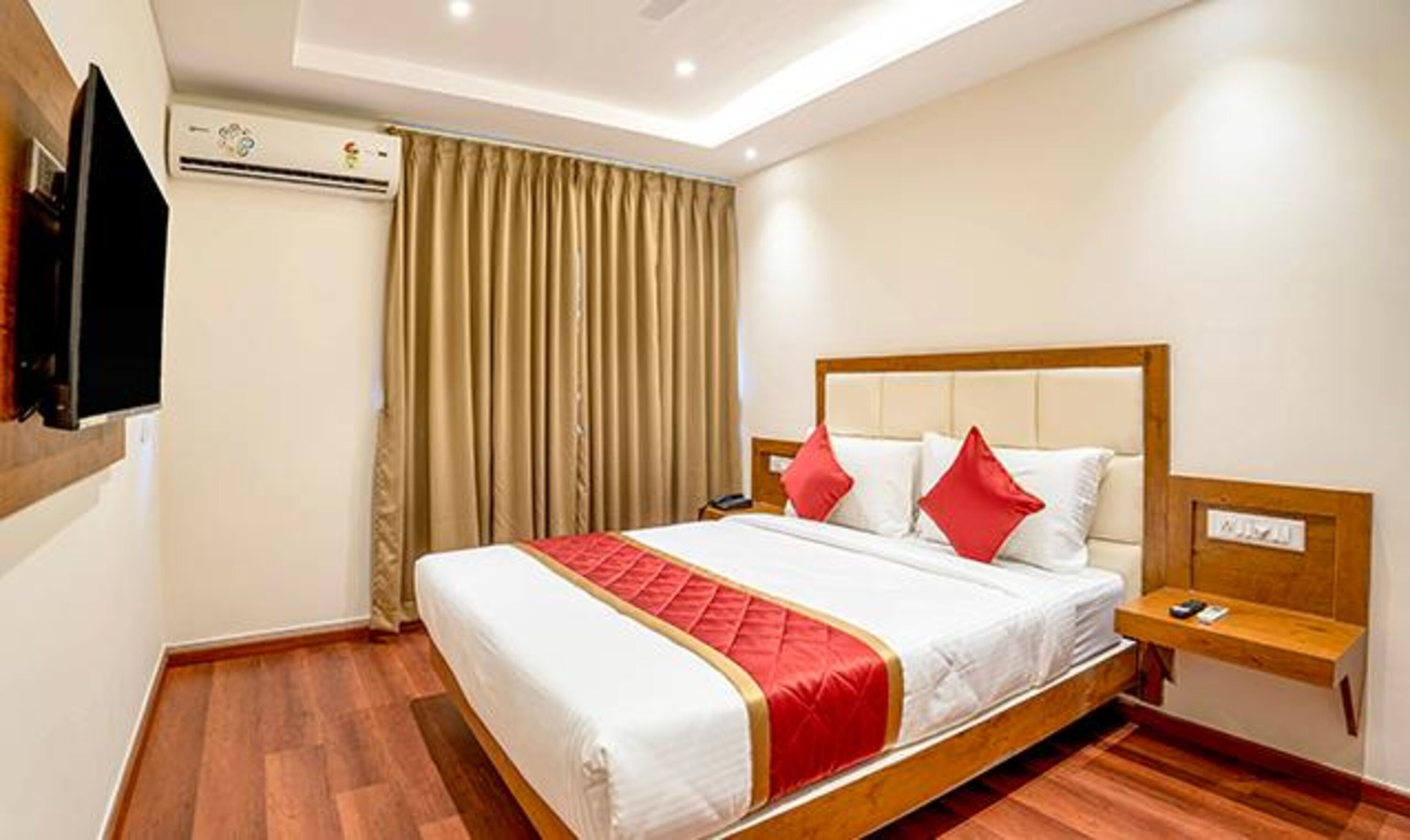 A hotel room with a bed, a bedside table, a TV mounted on the wall, and a window with curtains.