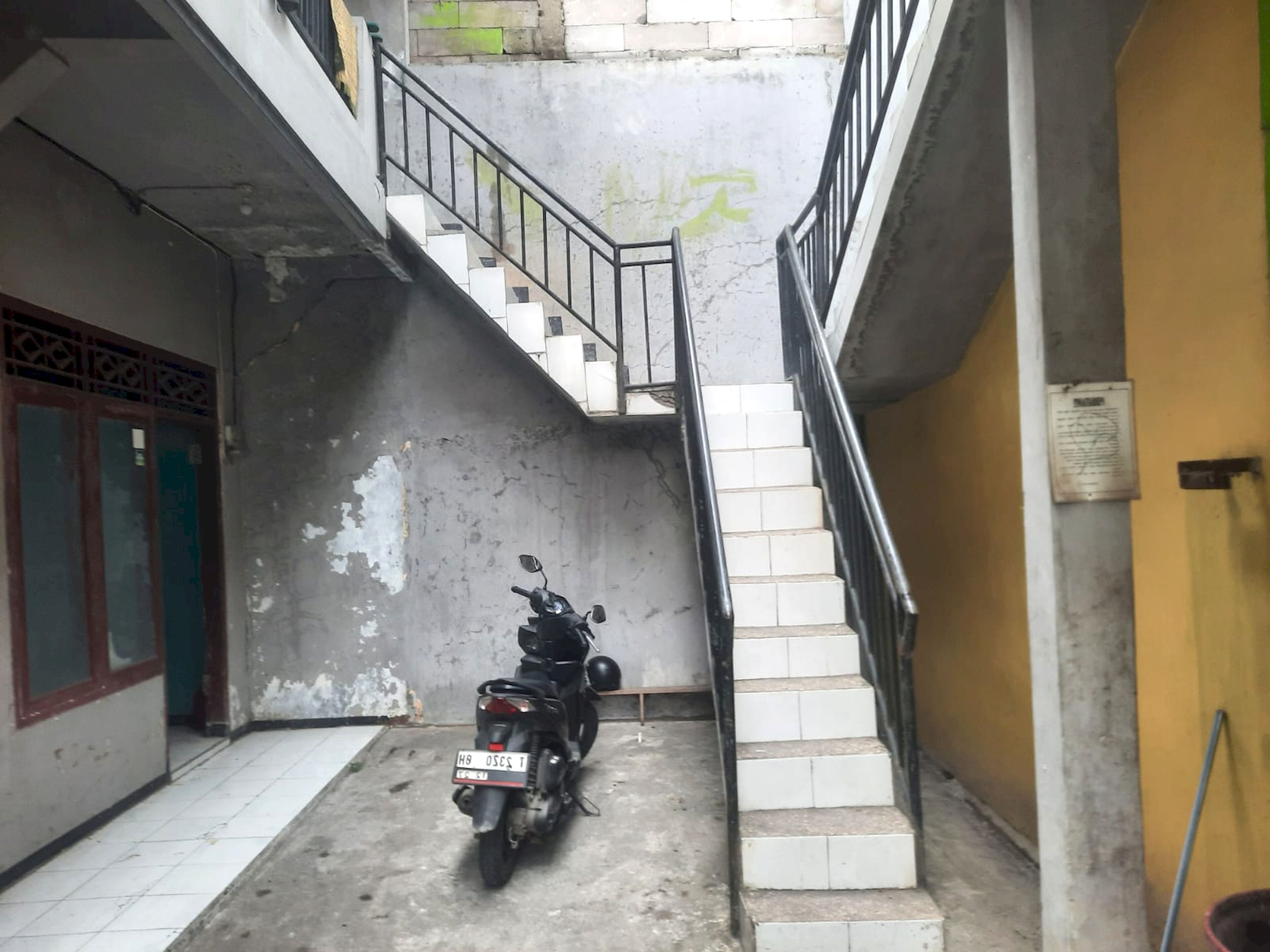 A narrow alleyway with a motorcycle parked at the bottom, a staircase leading upwards, and a door with a decorative grille on the left.