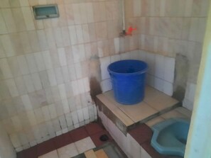 A small bathroom with a blue bucket, a toilet, and a tiled floor. - OYO Life  93023 Kost Bulan Karawang (Karawang)