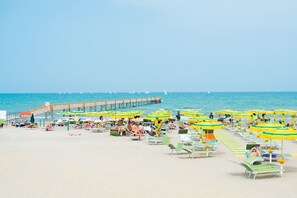 Uma praia com fileiras de espreguiçadeiras e guarda-sóis, um cais que se estende até o mar, e veleiros ao fundo.