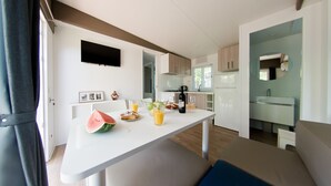A modern kitchen with a white countertop, a refrigerator, and a sink. There is a dining table with a chair, and a large window providing natural light. - Comfortable, fully equipped, 2-bedroom, 2-bathroom mobile home, Cesenatico beach (Cesenatico)