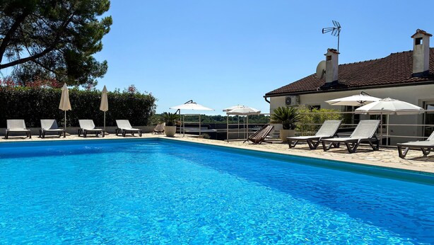 Una piscina con sillones de playa y sombrillas, rodeada por un edificio con techo de tejas.