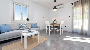 Un salón moderno con sofá, una mesita y una zona de comedor con sillas. Se aprecia una ventana con vistas, un equipo de aire acondicionado y una lámpara de techo.