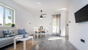 Una sala de estar moderna con sofá, mesa de centro, mesa de comedor y sillas. Hay un televisor montado en la pared y una ventana con cortinas.
