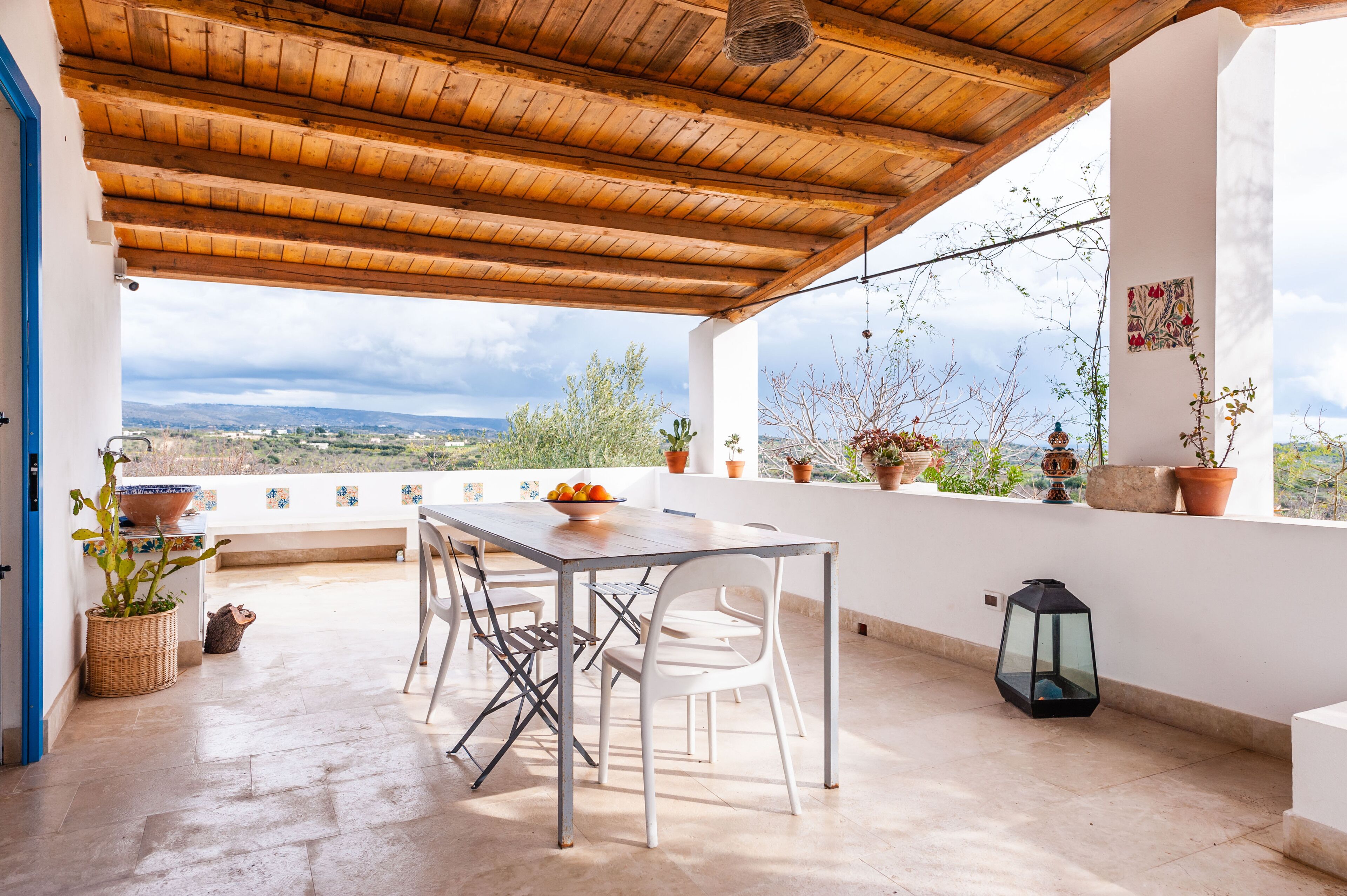 An outdoor patio with a wooden ceiling, a table with chairs, potted plants, and a view of the landscape.