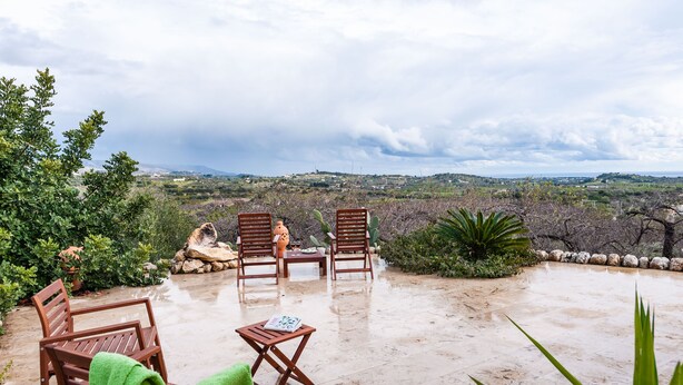 Un espace extérieur aménagé avec des chaises et une table en bois, entouré de plantes et de rochers, et offrant une vue sur un paysage vallonné.