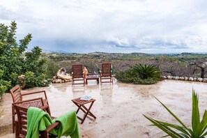 Un espace extérieur aménagé avec des chaises et une table en bois, entouré de plantes et de rochers, et offrant une vue sur un paysage vallonné.