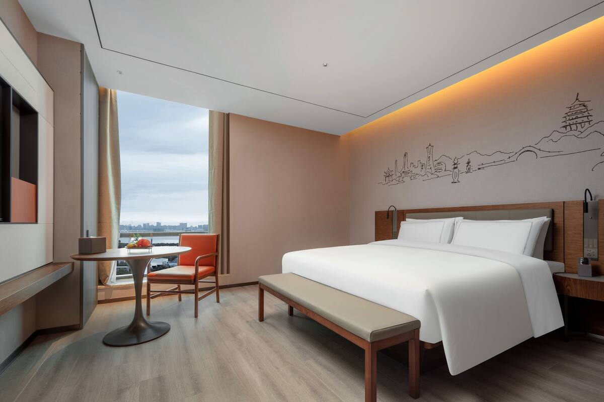 Ein modernes Hotelzimmer mit einem großen Bett, einem Schreibtisch mit Stuhl, einer Bank und Blick auf die Skyline durch große Fenster.