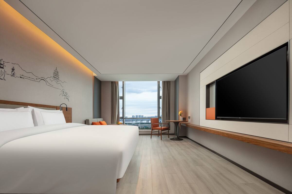 Ein modernes Hotelzimmer mit einem großen Bett, einem Schreibtisch mit Stuhl, einem Flachbildfernseher und Blick auf die Stadt durch große Fenster.