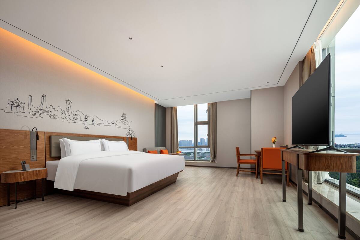 Ein modernes Hotelzimmer mit einem großen Bett, einem Schreibtisch mit Fernseher und Blick auf die Stadt.